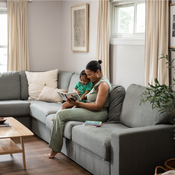 A woman in a matching sage green loungewear set sits barefoot on a gray sectional sofa, reading a book while holding a child. The scene is set in a bright, modern living room with beige curtains, a wooden coffee table, and a houseplant.