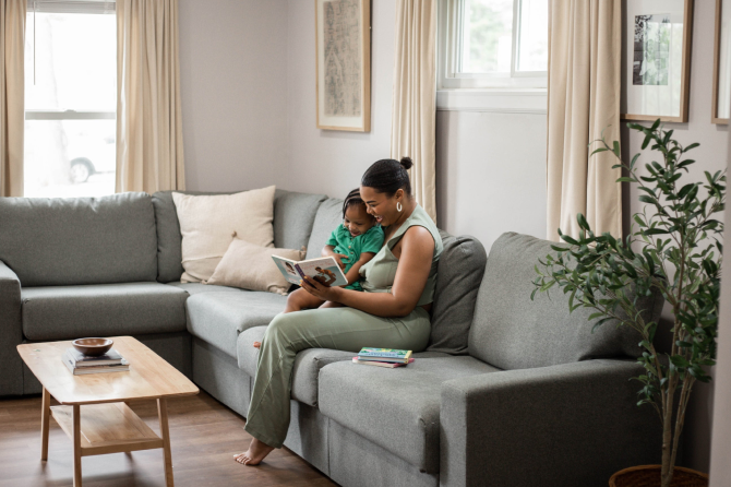 A woman and a child sit on a large gray sectional sofa in a bright living room, reading a book. The scene includes a light wood coffee table, beige curtains, framed wall art, and a potted plant.