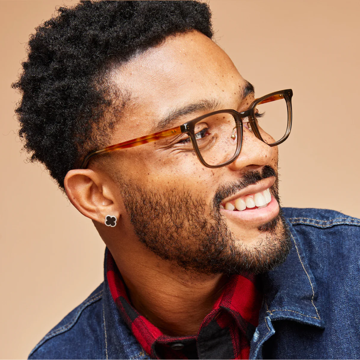 A close-up portrait of a smiling man with short curly black hair, wearing tortoiseshell eyeglasses with a dark brown frame, a red and black plaid shirt, and a blue denim jacket, set against a solid beige background.